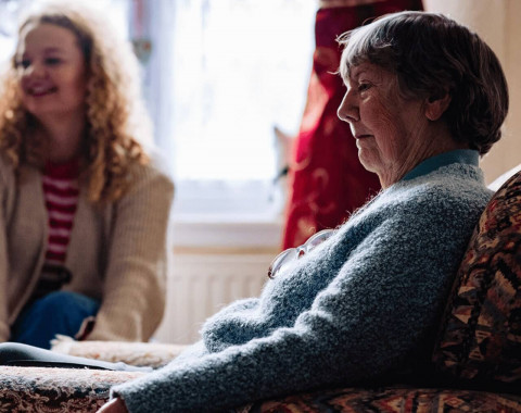 An older woman with anxiety is sitting looking anxious. A smiling younger woman sits behind her looking away from her.