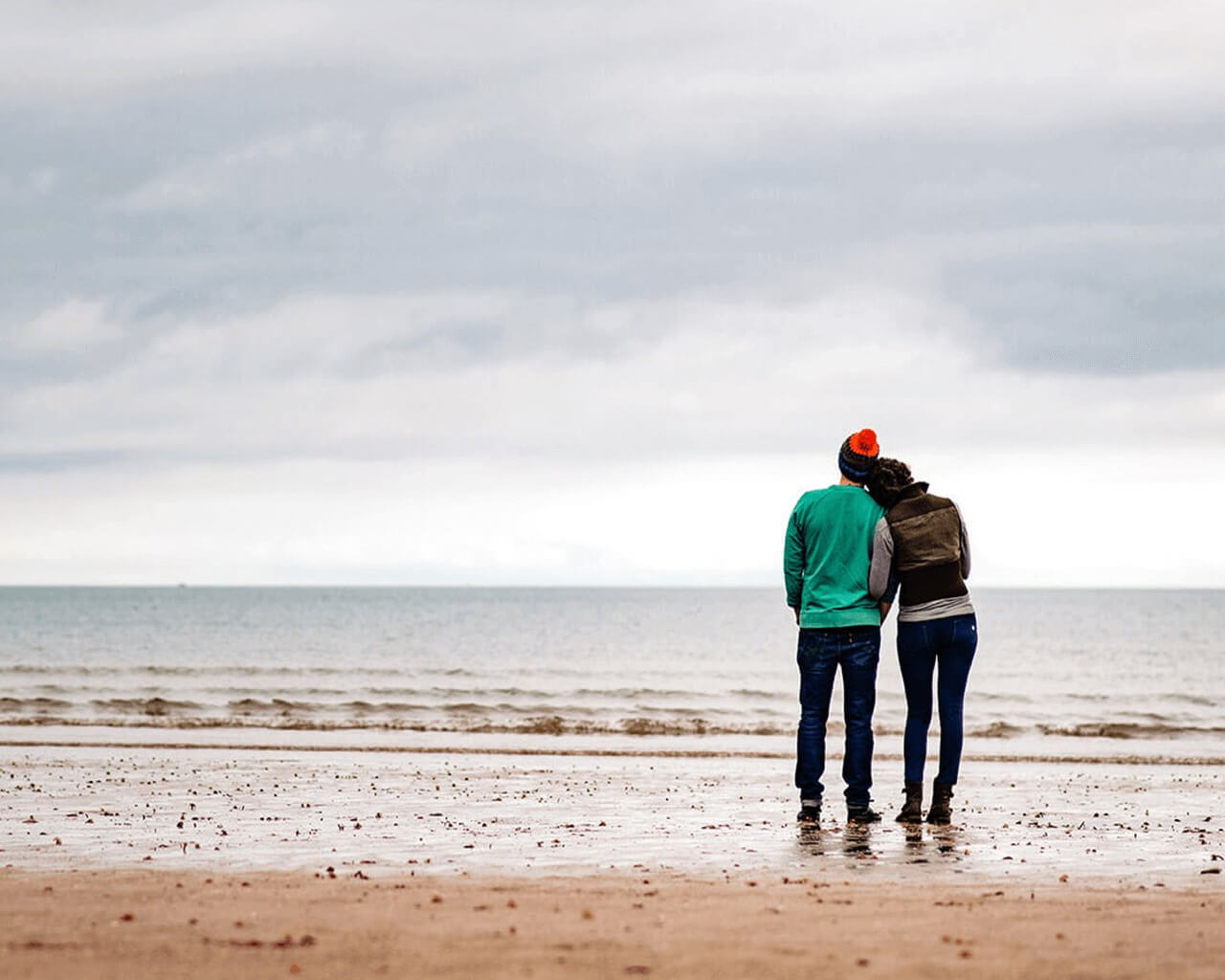 A tired couple standing on a beach looking out to sea. The woman is resting her head on the man's shoulder.