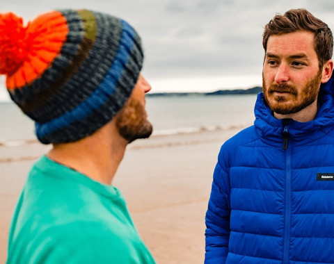 Two men are stood on a beach on a grey day. They look like they are having a serious conversation based on their expressions.