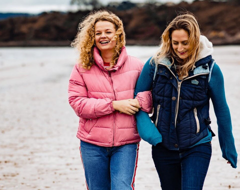 Two women are walking along the beach together with their arms linked, smiling.
