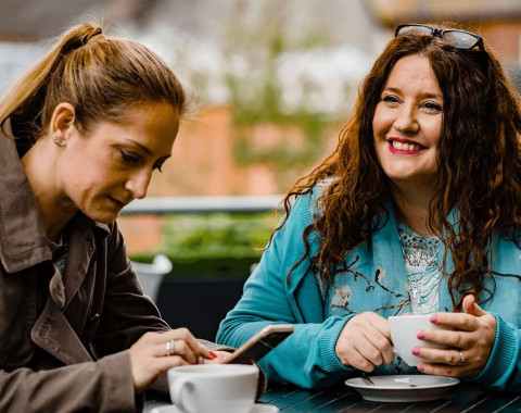 Two women are sitting having coffee together outside. One of the women is looking at something on her mobile phone.