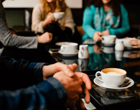 A group of people are sitting having coffee. A man has his hands clasped looking anxious and hasn't started his coffee.