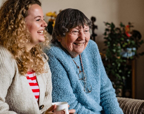 Two women are sitting together on a sofa laughing and smiling.