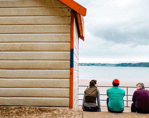 Three people sitting next to a beach hut looking out to sea on a cloudy day.