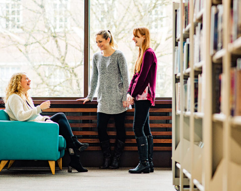 Three people are talking in a library by a window. One is sat down in a chair laughing and other two are standing.