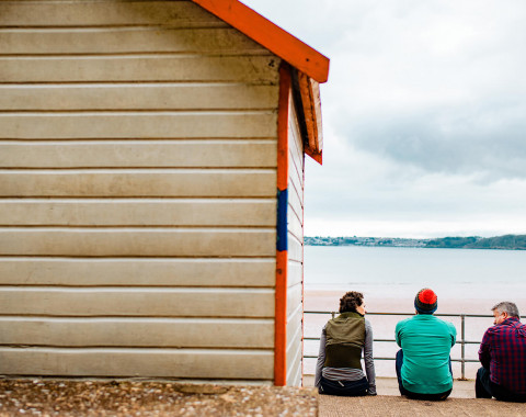 Three people are sat at the beach next to the right hand side of a beach hut facing the sea on a cloudy day.