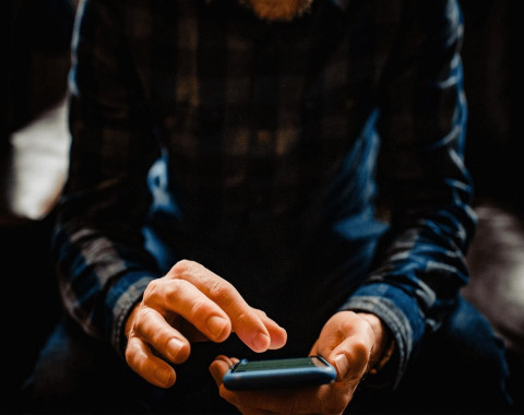 A man is sitting in a dimly lit room. He is dressed in black and is using a mobile phone.